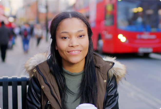 A young woman seated on street bench enjoying coffee as red bus travels past in background