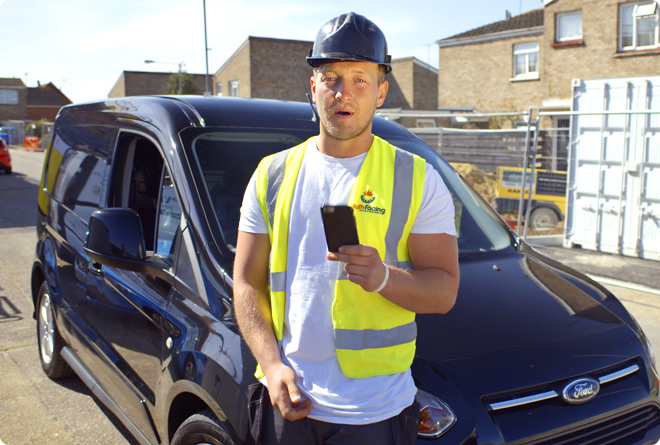 A young tradesman standing in front of his van holding his mobile phone at a building site